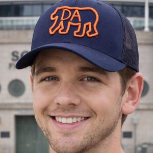 May include: Navy blue trucker hat with an orange outline of a bear and the letters "PA". The hat has a curved brim and a mesh back. A stylish accessory for outdoor activities or casual wear.