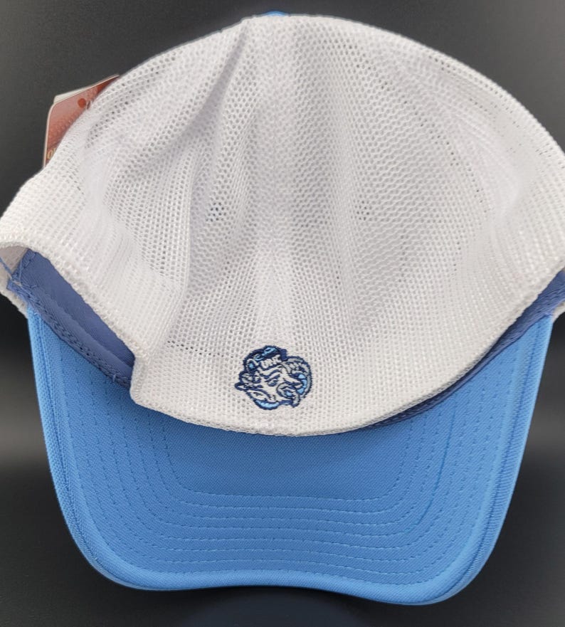 May include: A white and light blue baseball cap with a mesh back. The front of the cap features a blue and white embroidered logo of a cartoon creature with the text "UNC" above it.