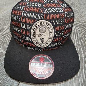 May include: Black and red Guinness Extra Stout baseball cap with a black brim. The cap features the Guinness logo patch and the words "Guinness" repeated across the top. The cap also has a "American Needle" patch.