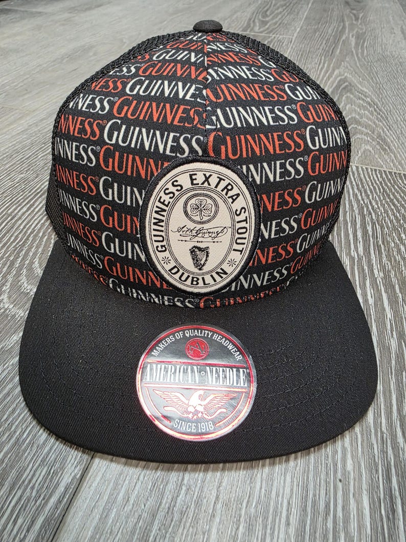 May include: Black and red Guinness Extra Stout baseball cap with a black brim. The cap features the Guinness logo patch and the words "Guinness" repeated across the top. The cap also has a "American Needle" patch.