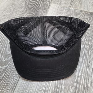 May include: Black trucker hat with a mesh back and a curved brim. The hat has a snap closure for adjustable sizing. The underside of the brim is a light pink color. The hat is sitting on a wooden surface.