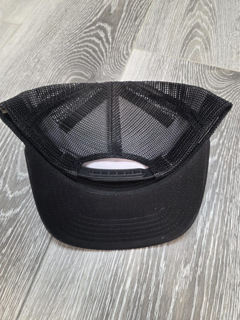 May include: Black trucker hat with a mesh back and a curved brim. The hat has a snap closure for adjustable sizing. The underside of the brim is a light pink color. The hat is sitting on a wooden surface.