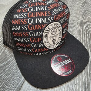 May include: Black and mesh-backed baseball cap featuring the Guinness logo in red, white, and black. The cap has a flat brim and a circular patch with the Guinness Extra Stout logo. The American Needle logo is also visible.