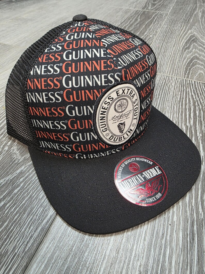 May include: Black and mesh-backed baseball cap featuring the Guinness logo in red, white, and black. The cap has a flat brim and a circular patch with the Guinness Extra Stout logo. The American Needle logo is also visible.