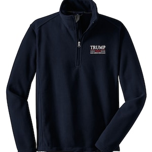 May include: Navy blue fleece pullover with a quarter-zip closure and a stand-up collar. The chest features the text "TRUMP 2024" in white and red, with "MAKE AMERICA GREAT AGAIN" below it. The long-sleeved top is made of a soft, warm material.