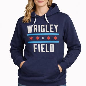 May include: Navy blue hooded sweatshirt with the text "WRIGLEY FIELD" in white block letters. The design includes blue stripes and red stars. The sweatshirt has a front pocket and a white drawstring hood.