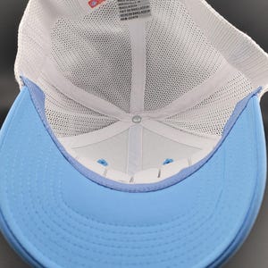 May include: The inside of a white and light blue baseball cap. The cap is made of mesh and has a white label that reads "American Needle" and "Made in Bangladesh".