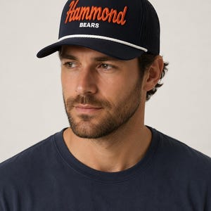 May include: A navy blue baseball cap featuring a white rope detail across the brim. The front displays the word Hammond in orange script embroidery above the word BEARS in white block lettering. The back of the cap is made of navy mesh fabric.