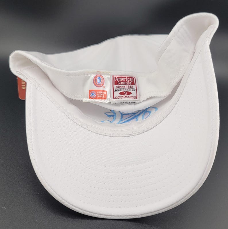 May include: A white baseball cap with a curved brim, featuring a light blue embroidered design on the underside. The cap has a tag that reads "American Needle since 1918 TECH/STRETCH".