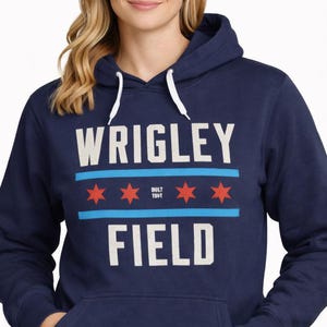 May include: Navy blue hooded sweatshirt with the text "WRIGLEY FIELD" in white block letters. The design includes a light blue stripe with three red stars, and the text "BUILT 1914". The sweatshirt has a front pocket and white drawstrings.