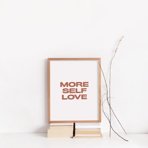 May include: A framed print with the text "MORE SELF LOVE" in a brown font on a white background. The frame is a light wood color and the print is sitting on a stack of books.