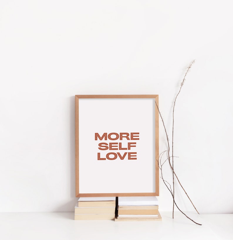 May include: A framed print with the text "MORE SELF LOVE" in a brown font on a white background. The frame is a light wood color and the print is sitting on a stack of books.