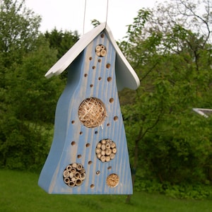 Può includere: Una casetta di legno blu per insetti con un tetto bianco e diversi fori per permettere agli insetti di nidificare. La casetta è appesa a una corda.