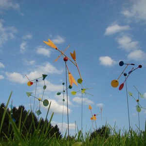 May include: A field of colorful, abstract metal sculptures with round and triangular shapes. The sculptures are attached to long metal poles and are spinning in the wind. The sculptures are against a blue sky with white clouds.