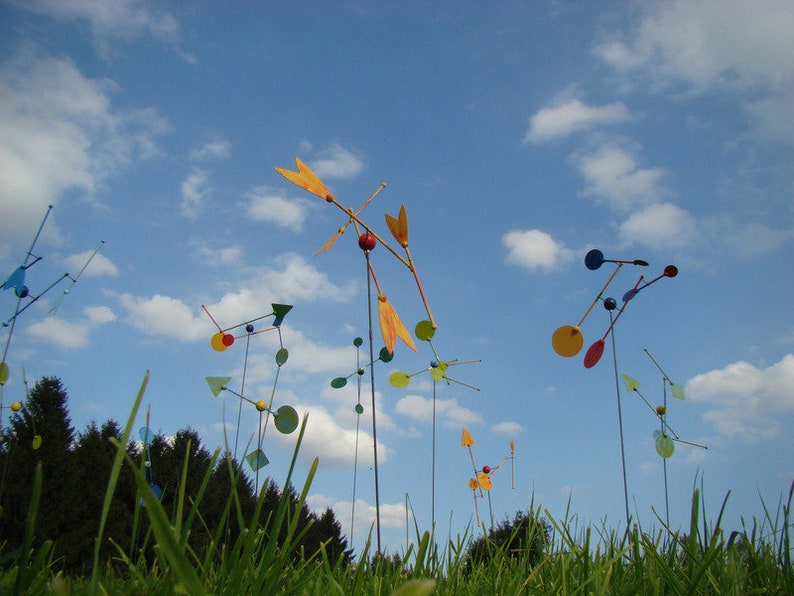 May include: A field of colorful, abstract metal sculptures with round and triangular shapes. The sculptures are attached to long metal poles and are spinning in the wind. The sculptures are against a blue sky with white clouds.