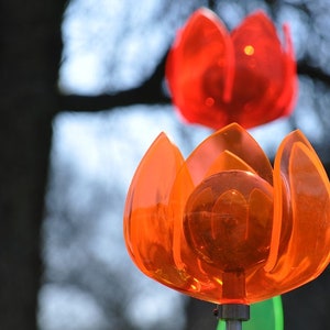 Puede incluir: Dos molinetes de viento de plástico en forma de flor de color naranja y rojo con un centro redondo y transparente. Los molinetes de viento están colgados de una varilla de metal.