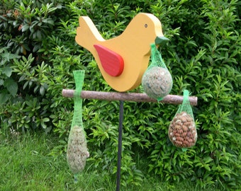 Pode incluir: Um comedouro de pássaros de madeira com uma figura de pássaro amarelo segurando um saco de malha de sementes para pássaros. Duas bolsas de malha adicionais de sementes para pássaros penduram de um galho abaixo do pássaro. O comedouro é montado em um poste de metal preto.
