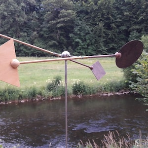 Peut inclure: Un moulin à vent en bois avec quatre pales de formes différentes. Les pales sont un triangle, un cercle, un ovale et un rectangle. Le moulin à vent est monté sur un poteau en métal et se trouve dans une zone herbeuse près d'une rivière.