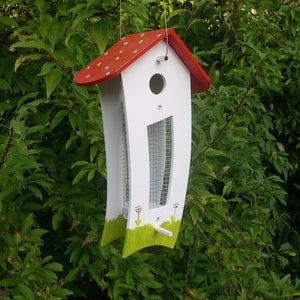 May include: White bird feeder with a red roof and white polka dots. The feeder has a green grass design at the bottom and a mesh opening for birds to access the food.