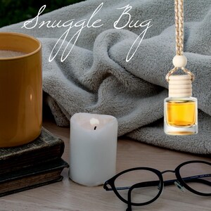 May include: A white candle, a mug of coffee, a gray throw blanket, and a glass bottle with a wooden cap hanging from a brown cord. The bottle is filled with a golden liquid. The text "Snuggle Bug" is written in white cursive on the image.