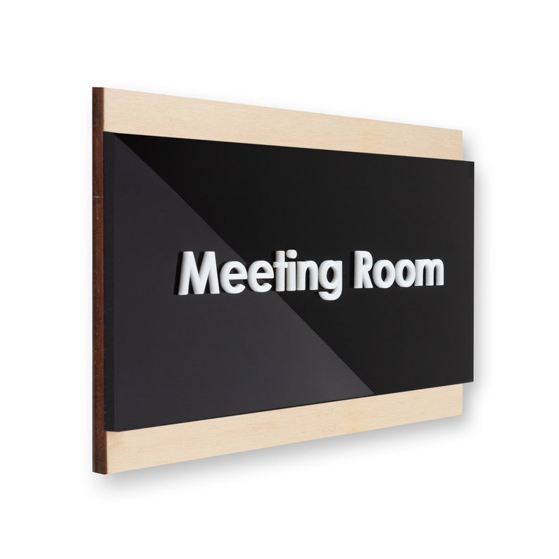 May include: A black sign with white raised lettering that reads "Meeting Room" is framed with a light wood border.