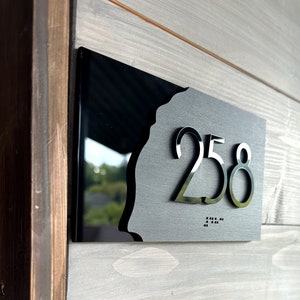 Puede incluir: Placa de número de casa negra con el número 258 en letras blancas en relieve. La placa tiene un fondo negro con un borde blanco en relieve irregular. La placa también tiene letras en braille debajo del número.