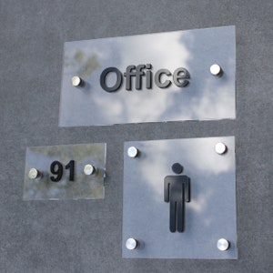 Puede incluir: Tres letreros de vidrio transparente con letras negras y detalles plateados. El letrero superior dice "Office". El letrero del medio dice "91". El letrero inferior tiene una silueta negra de una persona.