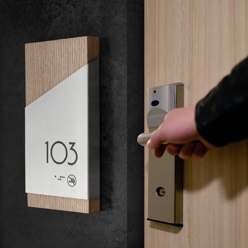 May include: A wooden hotel room sign with a white panel displaying the number "103". The sign has a no smoking symbol and braille dots.