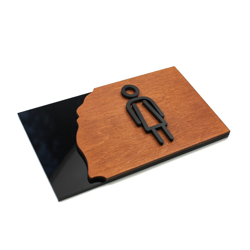 May include: A black and brown wooden sign with a stylized female figure icon. The sign has a black background with a brown wooden overlay.
