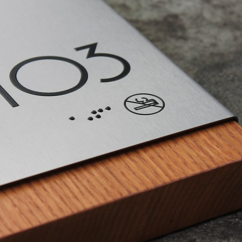 May include: A silver metal sign with the number 103 and a no smoking symbol. The sign is mounted on a wooden base.
