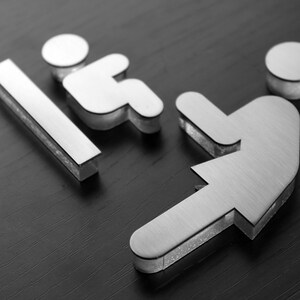 May include: A silver metal sign with two figures representing a restroom. The figure on the left is a stick figure sitting down, and the figure on the right is a stick figure standing up.