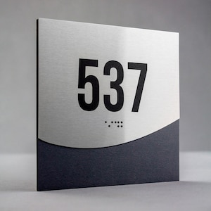 May include: A silver and black room number sign with the number 537 in black lettering. The sign has a braille number underneath the number 537.