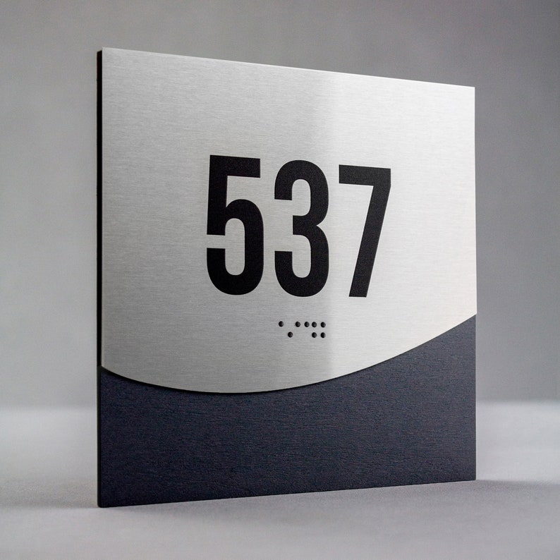 May include: A silver and black room number sign with the number 537 in black lettering. The sign has a braille number underneath the number 537.