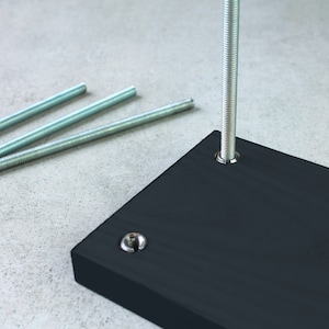 May include: A black wooden base with a silver metal rod extending upwards. Three silver metal rods are lying on a gray surface.
