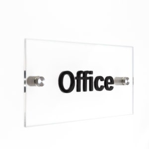 May include: A clear acrylic sign with black raised lettering that reads "Office". The sign is mounted to a white wall with two silver screws.