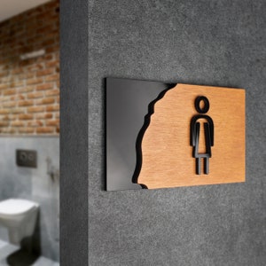 Puede incluir: Un letrero negro y marrón con un icono estilizado de una figura femenina. El letrero tiene un borde ondulado y está montado en una pared gris.