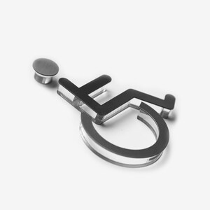 May include: A silver wheelchair accessibility symbol with a black outline. The symbol is made of acrylic and has a small silver pin on the back.