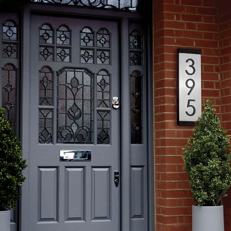 May include: A gray front door with a glass panel and a silver mailbox. A house number plaque with the number 395 is mounted on the brick wall next to the door.