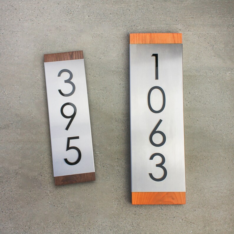 May include: Two vertical house number signs with black numbers on a silver background and a wood frame. The numbers on the left sign are 395 and the numbers on the right sign are 1063.
