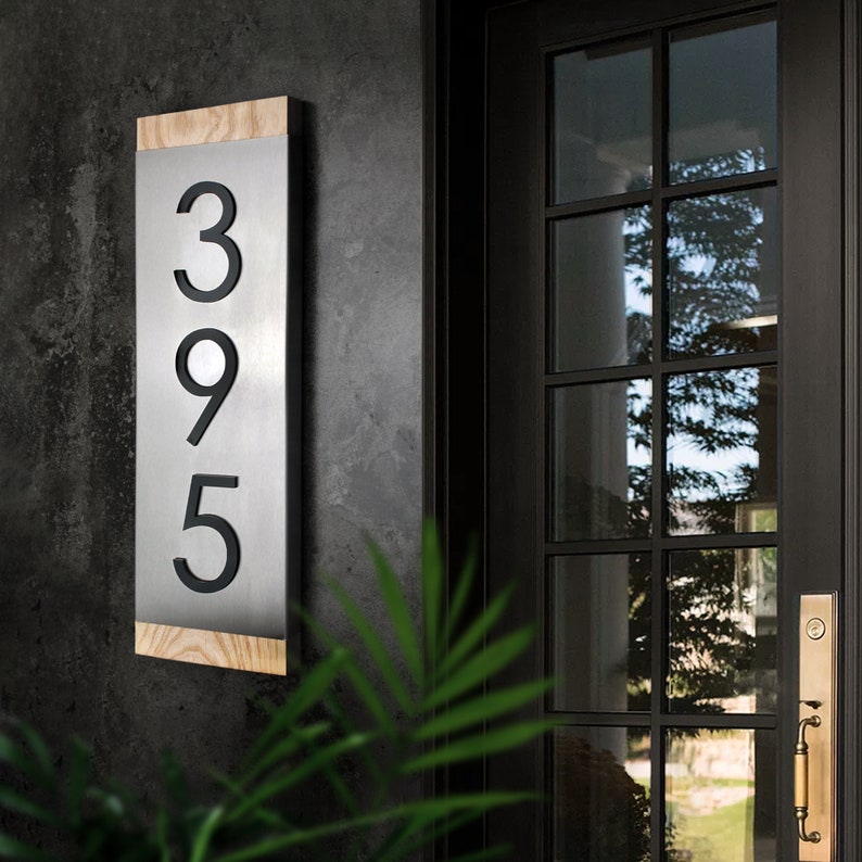 May include: A modern house number sign with the numbers 395 displayed in black on a silver metal background. The sign is mounted on a wooden frame.