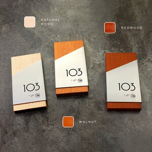 May include: Three wooden house number signs with a silver metal plate. The signs are in natural wood, walnut, and redwood. Each sign has the number 103 on the metal plate.