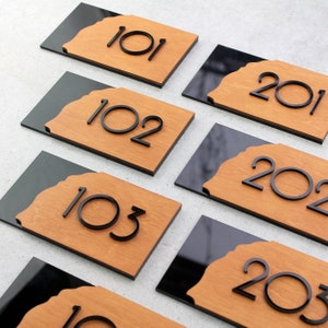 Puede incluir: Cinco placas de números de casa de madera con fondos negros. Los números son 101, 102, 103, 201 y 202.