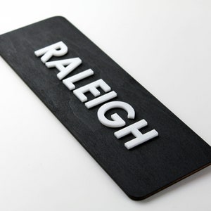 Puede incluir: Un letrero de madera negro con el nombre "RALEIGH" en letras blancas en relieve.