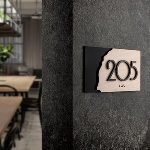 Puede incluir: Un letrero negro y de madera con el número 205 en letras negras. El letrero tiene un borde ondulado y está montado en una pared gris.