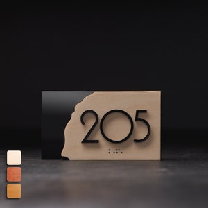 Puede incluir: Un letrero rectangular de madera con el número "205" en negro, con un acento negro en el lado izquierdo. El letrero incluye puntos en braille debajo de los números. El fondo es oscuro.