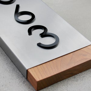 May include: A silver metal house number plaque with black acrylic numbers "63" mounted on a wooden base.