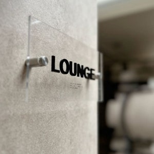Puede incluir: Un letrero de acrílico transparente con la palabra "LOUNGE" en letras negras. El letrero está montado en una pared con un soporte plateado.