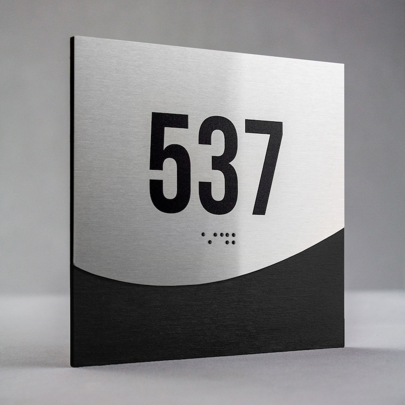 May include: A silver and black room number sign with the number 537 in black lettering. The sign has braille dots below the number.