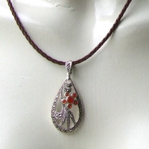 Puede incluir: Un collar de plata con un colgante en forma de lágrima que presenta una flor roja y un intrincado trabajo de filigrana. El collar está en un cordón de cuero marrón.