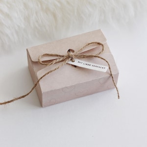 Könnte beinhalten: Eine kleine, hellrosa Geschenkbox, gebunden mit Bindfaden und einer Schleife. Die Box hat ein Etikett mit der Aufschrift "MIT LIEBE VERPACKT". Die Box ist mit herzförmigen Mustern verziert und liegt auf einer weißen Oberfläche.